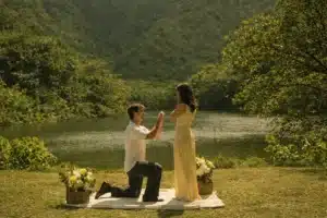 idea | Romantic outdoor proposal by a lakeside, with a man kneeling on a blanket holding a ring box while a woman in a soft yellow dress stands surprised, surrounded by flower baskets, lush green hills, and warm golden sunlight in a serene 16:9 landscape setting.