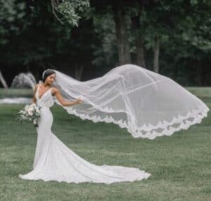 A bride in a fitted white lace gown stands outdoors on a grassy field, holding a bouquet of flowers, while her long embroidered veil billows dramatically in the wind behind her. bridal veils
