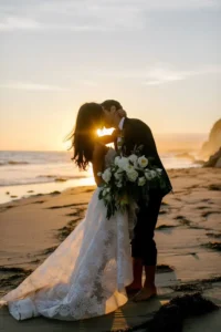 Beach Wedding | *Silhouette of a bride and groom standing under a floral arch on a beach at sunset, framed by tall palm trees, with guests seated on either side and the ocean glowing in the background.*