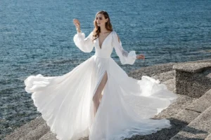 Personal style | Bride in a flowing white chiffon wedding gown with long sheer sleeves and a thigh-high slit, standing gracefully on stone steps beside the ocean. Her dress billows dramatically in the sea breeze as sunlight reflects off the blue water behind her, creating a soft, romantic coastal setti