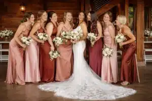 bridal retinue | Bride in a white lace gown standing at the centre, surrounded by a large bridal party of bridesmaids in coordinated blush and bronze satin dresses, all smiling and holding bouquets inside a rustic wooden wedding venue.