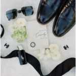 Groom's style guide - Black leather groom’s shoes, sunglasses, vow booklet, black ring, cufflinks, cologne bottle, black bow tie ribbon, and white flowers arranged flat on a light background.