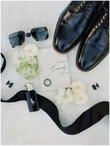 Groom's style guide - Black leather groom’s shoes, sunglasses, vow booklet, black ring, cufflinks, cologne bottle, black bow tie ribbon, and white flowers arranged flat on a light background.
