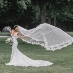 A bride in a fitted white lace gown stands outdoors on a grassy field, holding a bouquet of flowers, while her long embroidered veil billows dramatically in the wind behind her. bridal veils
