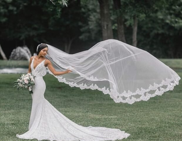A bride in a fitted white lace gown stands outdoors on a grassy field, holding a bouquet of flowers, while her long embroidered veil billows dramatically in the wind behind her. bridal veils