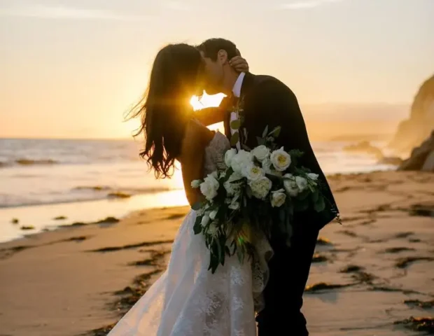 Beach Wedding | *Silhouette of a bride and groom standing under a floral arch on a beach at sunset, framed by tall palm trees, with guests seated on either side and the ocean glowing in the background.*