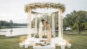 Poruwa Ceremony | Elegant Sri Lankan bride and groom performing a traditional Poruwa ceremony beneath a white floral canopy beside a serene lakeside setting, surrounded by symbolic ceremonial elements and lush greenery.